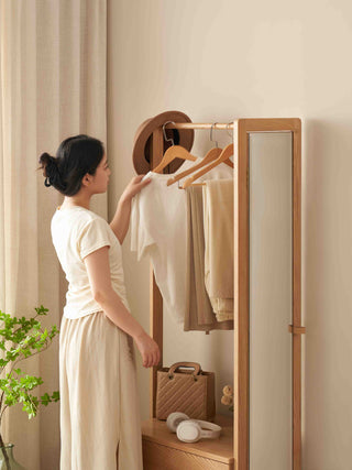 Woman in a white outfit standing in front of a wooden wardrobe with a mirror, holding a white shirt.