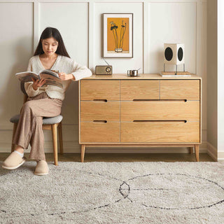 Woman reading a book in a room with a wooden dresser and decorative items.