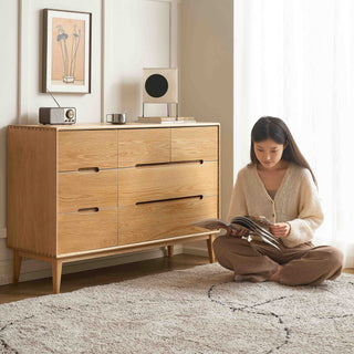 Woman reading a book in front of a solid wood chest of drawer in a bright room.