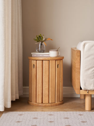 Wooden side table with a plant, books, and a cup in a room with a curtain and chair.