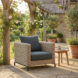 Woven outdoor chair with dark cushions on a patio with garden and pergola in the background