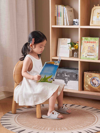 Young girl reading a book in a room with a wooden shelf and decorative items.