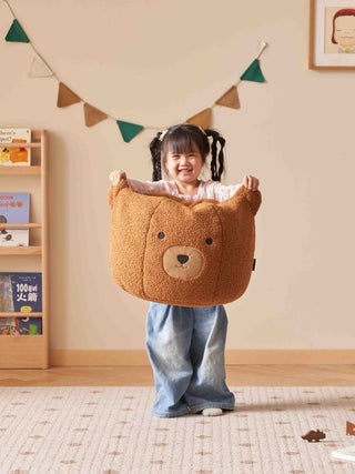 kids holding a brown bear-shaped bean bag chairs in a room with a bookshelf and colorful flags.