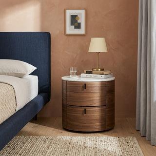 round bedside table with books and a lamp next to a bed in a bedroom.