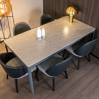 A modern dining table with a gray sintered stone top and black aluminum frame, accompanied by four black chairs with curved legs. There is a lamp with a gold-colored shade on the table, and two white bottles on top of the table.